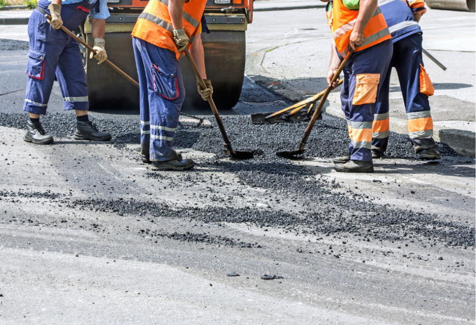 Road construction crew in high-visibility orange vests and blue workwear using rakes and shovels to spread fresh asphalt behind a steamroller.